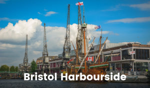 A historic tall ship is moored at Bristol Harbourside. Its timber construction gleams in the sun and flags from England and Canada fly from its masts. There are old cranes on the wharf and a large building set back from the water.