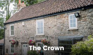 An old stone building with mostly white painted windows and door, and a black-framed bay window, with colourful flower baskets hang from the wall. There is a small pub garden out front with white wrought iron tables and chairs dotted about. A small black sign with ‘The Crown’ written on it in gold lettering hangs on the wall.