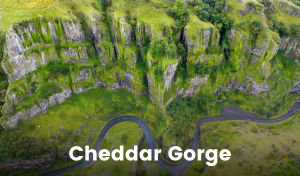 An overhead view of the iconic s-bend of the road travelling through Cheddar Gorge. Towering cliffs streaked with trees and foliage reach up to the sky, and footpaths are traced through the grass along the top of the gorge.