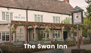 A large building, half brick and half timber framed, with white plaster, blue-grey woodwork and mullion windows. The paved patio is filled with picnic benches and umbrellas, and a large sign hanging on the pergola entrance to the pub says ‘Woodborough Inn’
