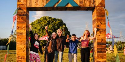 Group of smiling kids and adults posing under a wooden entrance arch at Mendip Basecamp during a summer family adventure holiday filled with campfires, music, and outdoor fun